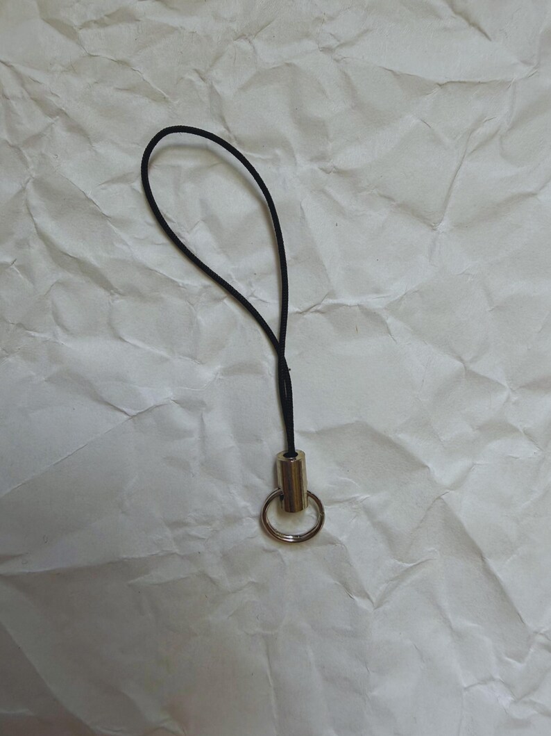 May include: A black lanyard with a gold-colored metal ring and barrel. The lanyard is looped and attached to the metal barrel. The background is a crumpled white paper.