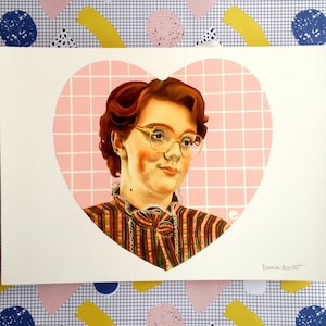 Puede incluir: Una impresión de retrato de una mujer pelirroja con gafas, enmarcada en un corazón rosa. Lleva una camisa a rayas. El fondo es un patrón de cuadrícula. La impresión está sobre papel rectangular blanco. Firmado "Emma Mount".