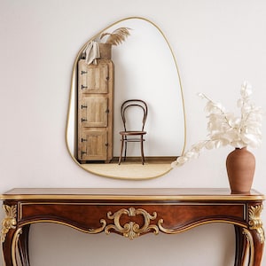 May include: An irregularly shaped gold-framed mirror reflects a wooden cabinet, chair, and vase with white flowers. The mirror hangs above a dark wood console table with gold accents. The scene evokes a warm, inviting aesthetic.