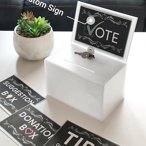 Peut inclure: Une boîte de scrutin rectangulaire en acrylique blanc avec serrure et clé, dotée d'une fente sur le dessus. La boîte comprend un porte-affiche amovible affichant le texte VOTE avec une coche. Des panneaux noirs supplémentaires avec du texte blanc indiquent SUGGESTION BOX, DONATION BOX et TIP$ Greatly Appreciated.