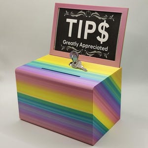 Op de afbeelding: Een regenboogkleurige houten fooienbox met een gleuf aan de bovenkant. De doos heeft een roze omlijst bord met de tekst "TIPS $ Greatly Appreciated". Een sleutel en een slot zijn zichtbaar op de bovenkant.