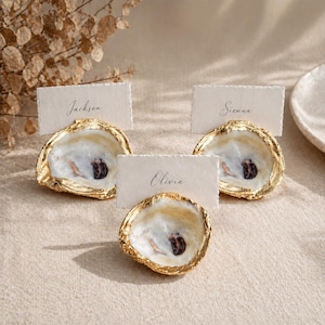 May include: Three place card holders made from natural oyster shells with gilded gold edges. Each shell supports a rectangular, off-white textured paper card with a deckled edge. The names Jackson, Sienna, and Olivia are printed in black script.