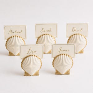 Könnte beinhalten: Fünf Platzkartenhalter in Form von Muscheln. Jeder Halter ist cremefarben mit goldenen Akzenten und hält eine cremefarbene Karte mit einem Namen in goldener Schrift: Michael, Sarah, David, Lisa und James. Sie stehen auf einer weißen Oberfläche.