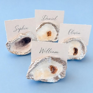 May include: Four place card holders made from oyster shells with silver-painted edges. Each shell holds a rectangular off-white card with a deckled edge. The cards feature the printed names Sophia, Daniel, Olivia, and William in black script.