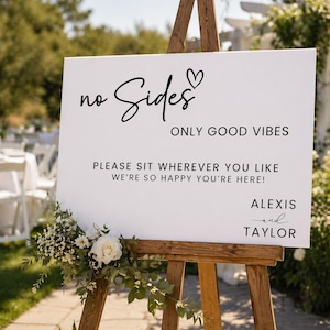 May include: A white wedding sign on a wooden easel with the words "no Sides" and "ONLY GOOD VIBES". The sign also reads "PLEASE SIT WHEREVER YOU LIKE, WE'RE SO HAPPY YOU'RE HERE!" and is signed "Alexis and Taylor". The sign is decorated with flowers.