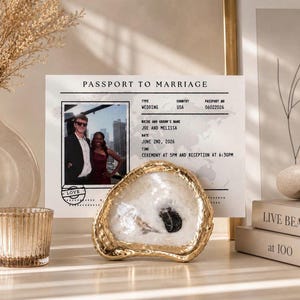 May include: A decorative gold-edged oyster shell dish with a black stone center sits in front of a wedding photo and a "Passport to Marriage" document. The document includes the names Joe and Melissa, and the date June 2nd, 2026.