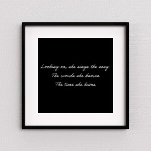 May include: A framed print with a black background and white text. The text reads, "Looking on, she sings the song, The words she knows, The tune she hums." The frame is black and the print is displayed on a white wall.