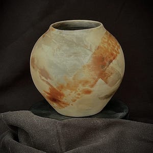 May include: A handcrafted ceramic vase with a rounded body and a wide opening. The vase features a blend of earthy tones, including beige, brown, and gray, creating a marbled effect. The vase is displayed on a dark surface against a dark background.