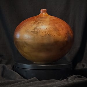 May include: A round, bulbous wooden vessel with a smooth, polished surface featuring swirling patterns of warm amber, deep brown, and reddish-orange tones. The object has a narrow, short neck at the top and a rounded body that tapers toward the base.