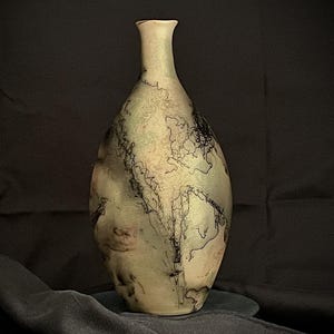 May include: A handcrafted, decorative vase with a unique, organic design. The vase features a light green and beige color palette with intricate black, lightning-like patterns. The elongated shape and narrow neck add to its elegant aesthetic. A striking piece for home decor.