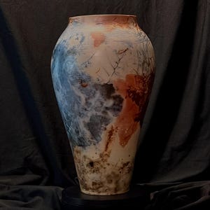 May include: A tall ceramic vase with a tapered base and a wide, rounded body. The surface features a mottled finish with blended patches of cream, deep blue, and burnt orange, accented by dark, intricate, web-like crackle patterns throughout.