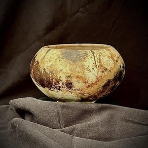 May include: A round, handcrafted ceramic bowl with a wide opening. The bowl features a mottled pattern of brown and tan hues, with darker speckles and streaks. The interior is a lighter tan color. The bowl sits on a dark surface.