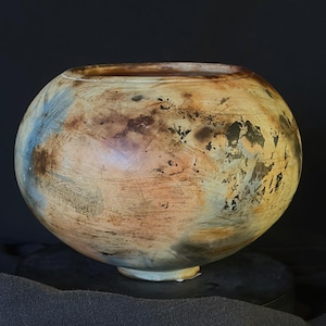 May include: A round, wide-mouthed bowl featuring a mottled surface with blended tones of tan, brown, cream, and soft blue. The exterior displays dark, irregular charred markings and fine linear textures across the curved, spherical body.