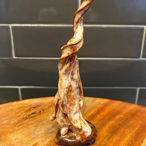 May include: A ceramic sculpture with a swirling, abstract design. The sculpture is mainly white with brown and orange accents, and it sits on a small, round, brown base. The piece has a textured appearance and a unique, artistic form.