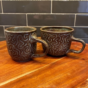 Puede incluir: Dos tazas de cerámica hechas a mano con un esmalte marrón y un patrón texturizado en espiral. Cada taza tiene un asa y un interior oscuro. Las tazas se exhiben sobre una superficie de madera.