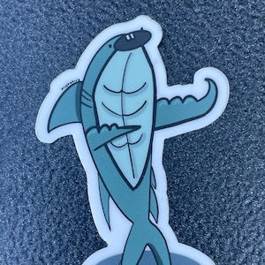 May include: A blue and white cartoon shark sticker with a black eye patch. The shark is standing on its tail with its fins out to the side. The sticker is on a blue background.