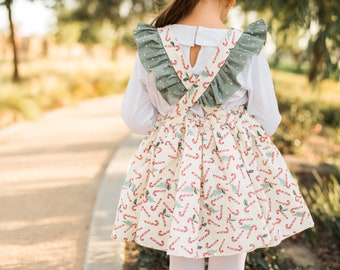 May include: A white dress with a candy cane and holly leaf print. The dress has a green ruffle trim and criss-cross straps in the back.