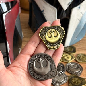 May include: Two metallic challenge coins featuring the Rebel Alliance symbol. One coin is finished in antique gold, while the other is in a dark gunmetal grey. Both coins display intricate circular engravings and Aurebesh-style text along the borders.