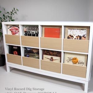 Puede incluir: Mueble blanco para guardar discos de vinilo con ocho compartimentos, cada uno con un cajón de roble claro. Muestra portadas de álbumes, incluyendo The Rolling Stones y Neil Young. El texto dice "Vinyl Record Dig Storage".