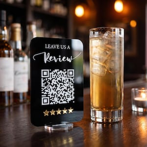 May include: A black rectangular acrylic sign with rounded corners featuring the white text LEAVE US A REVIEW above a white QR code and five gold star icons. The sign is supported by a clear circular base and stands upright.