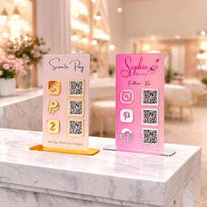 May include: Two acrylic display stands with QR codes. One is pink with the name "Sophia" and social media icons. The other is clear with "Scanto Pay" and payment symbols. Both have QR codes and are on a white surface.