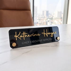 May include: A rectangular black acrylic desk nameplate featuring the gold script name Katherine Hayes and the white text CHIEF EXECUTIVE OFFICER. The sign is secured with two circular gold-toned metal standoffs on the front corners.