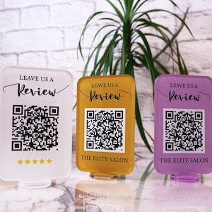 Op de afbeelding: Drie acrylstandaards met een QR-code en de tekst "Leave us a Review" op elk. De standaards zijn wit, geel en paars. De witte standaard heeft vijf gouden sterren onder de QR-code. De gele en paarse standaards hebben de tekst "The Elite Salon" onder de QR-code.