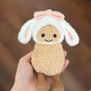 Może przedstawiać: Mała pluszowa owca z białym, puszystym futerkiem i brązowymi n&oacute;żkami. Owca ma na sobie biały, szydełkowy kapelusz z r&oacute;żowymi uszami kr&oacute;liczka i r&oacute;żową kokardką.