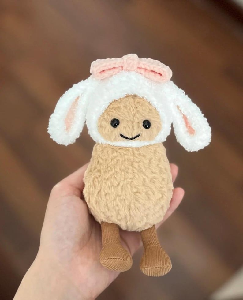 Może przedstawiać: Mała pluszowa owca z białym, puszystym futerkiem i brązowymi n&oacute;żkami. Owca ma na sobie biały, szydełkowy kapelusz z r&oacute;żowymi uszami kr&oacute;liczka i r&oacute;żową kokardką.