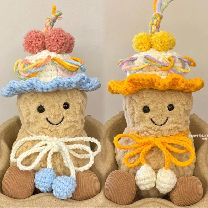 Puede incluir: Dos juguetes de peluche con forma de cacahuete que llevan sombreros y bufandas de ganchillo. El juguete de la izquierda lleva un sombrero azul con detalles de hilo rosa y amarillo y una bufanda blanca. El juguete de la derecha lleva un sombrero naranja con detalles de hilo amarillo y una bufanda amarilla.