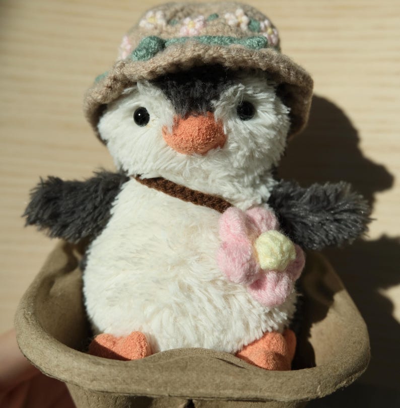K&ouml;nnte beinhalten: Ein wei&szlig;es Pl&uuml;schpinguin-Spielzeug, das einen braunen H&auml;kelhut mit Blumen und eine rosa Blume auf der Brust tr&auml;gt. Der Pinguin sitzt in einer braunen Papierschale.