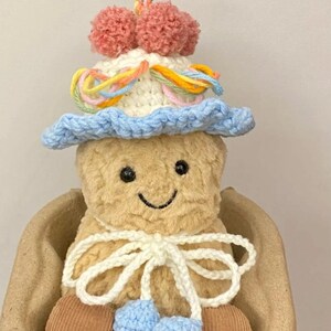 Puede incluir: Un juguete de peluche tejido a crochet que lleva un sombrero azul y blanco con detalles de hilo de colores y pompones. El juguete est&aacute; sentado en una cesta de mimbre marr&oacute;n.