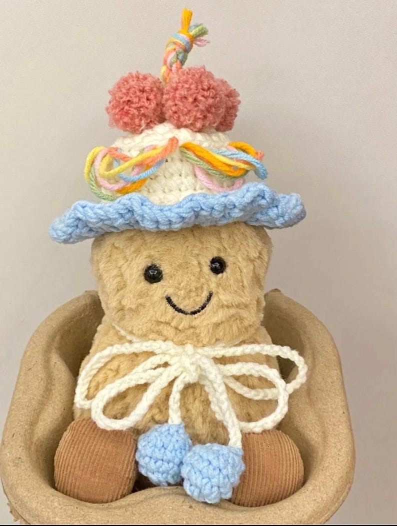 Puede incluir: Un juguete de peluche tejido a crochet que lleva un sombrero azul y blanco con detalles de hilo de colores y pompones. El juguete est&aacute; sentado en una cesta de mimbre marr&oacute;n.