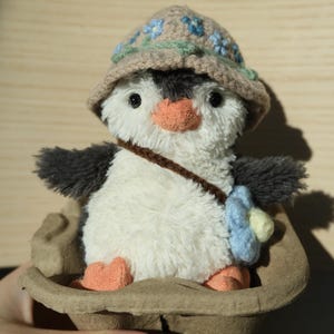 K&ouml;nnte beinhalten: Ein Pl&uuml;sch-Pinguin-Spielzeug, das einen braunen H&auml;kelhut mit blauen Blumen tr&auml;gt. Der Pinguin sitzt in einem braunen Papp-Eierkarton.