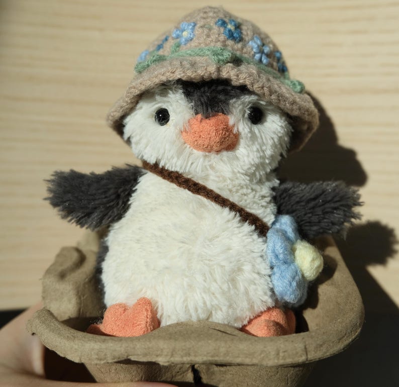 K&ouml;nnte beinhalten: Ein Pl&uuml;sch-Pinguin-Spielzeug, das einen braunen H&auml;kelhut mit blauen Blumen tr&auml;gt. Der Pinguin sitzt in einem braunen Papp-Eierkarton.