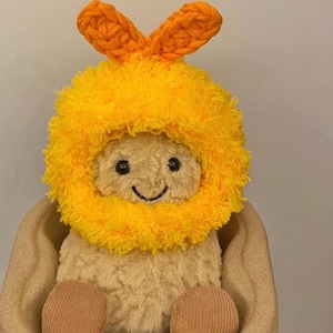 Könnte beinhalten: Ein Stofftier mit einem lächelnden Gesicht, das eine leuchtend gelbe, flauschige Kapuze mit orangefarbenen Hasenohren trägt. Das Spielzeug befindet sich in einem braunen Behälter mit Cordbeinen. Das Spielzeug ist weich und kuschelig.