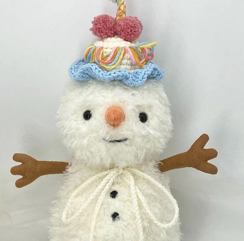 Puede incluir: Un mu&ntilde;eco de nieve de crochet blanco que lleva un gorro de crochet azul y amarillo con pompones rosas. El mu&ntilde;eco de nieve tiene brazos de lana marr&oacute;n y una bufanda de lana blanca.