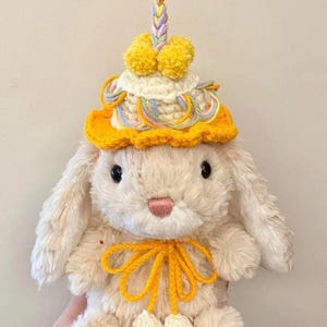 Puede incluir: Un conejito de peluche de color crema que lleva un gorro de crochet amarillo con un adorno de hilo trenzado de colores pastel y pompones. El conejito tambi&eacute;n tiene un lazo de crochet amarillo alrededor del cuello y pompones de crochet blancos en los pies.