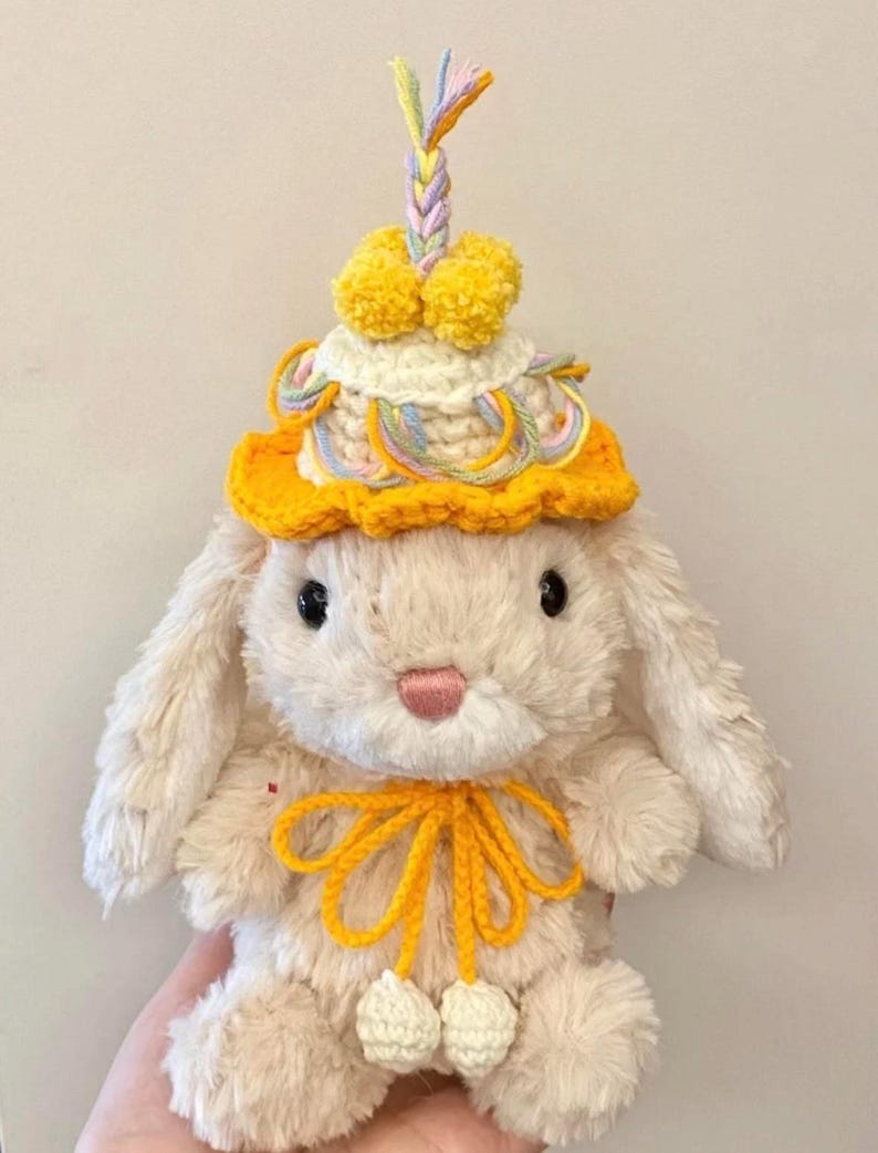 Puede incluir: Un conejito de peluche de color crema que lleva un gorro de crochet amarillo con un adorno de hilo trenzado de colores pastel y pompones. El conejito tambi&eacute;n tiene un lazo de crochet amarillo alrededor del cuello y pompones de crochet blancos en los pies.