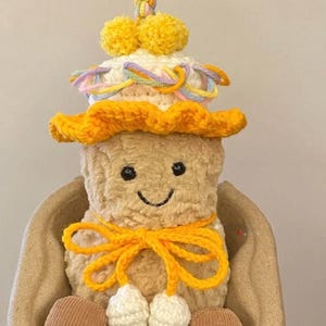 Puede incluir: Un juguete de peluche de cacahuete con un sombrero de ganchillo con pompones amarillos y un lazo amarillo. El cacahuete est&aacute; sentado en un cuenco de ganchillo marr&oacute;n.