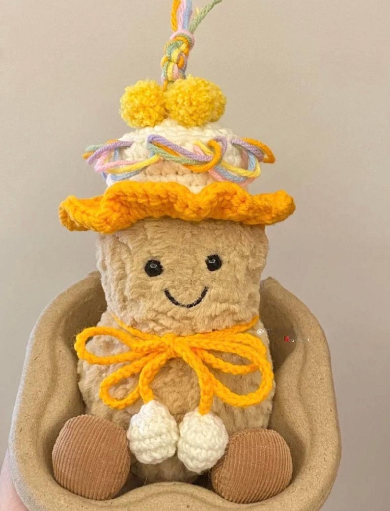 Puede incluir: Un juguete de peluche de cacahuete con un sombrero de ganchillo con pompones amarillos y un lazo amarillo. El cacahuete est&aacute; sentado en un cuenco de ganchillo marr&oacute;n.