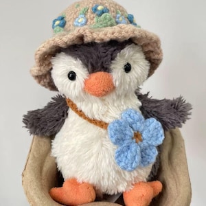 K&ouml;nnte beinhalten: Ein Pl&uuml;schpinguin mit einem geh&auml;kelten Hut mit blauen Blumen und einer blauen geh&auml;kelten Blume auf der Brust. Der Pinguin sitzt in einem braunen Papiernest.