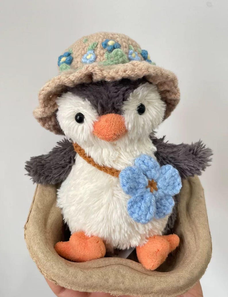 K&ouml;nnte beinhalten: Ein Pl&uuml;schpinguin mit einem geh&auml;kelten Hut mit blauen Blumen und einer blauen geh&auml;kelten Blume auf der Brust. Der Pinguin sitzt in einem braunen Papiernest.
