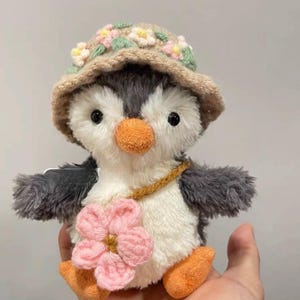K&ouml;nnte beinhalten: Ein Pl&uuml;schpinguin-Spielzeug, das einen braunen geh&auml;kelten Hut mit rosa und gr&uuml;nen Blumen tr&auml;gt. Der Pinguin hat einen wei&szlig;en K&ouml;rper, graue Fl&uuml;gel und eine rosa Blume, die an seiner Seite befestigt ist.