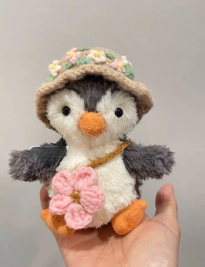 K&ouml;nnte beinhalten: Ein Pl&uuml;schpinguin-Spielzeug, das einen braunen geh&auml;kelten Hut mit rosa und gr&uuml;nen Blumen tr&auml;gt. Der Pinguin hat einen wei&szlig;en K&ouml;rper, graue Fl&uuml;gel und eine rosa Blume, die an seiner Seite befestigt ist.
