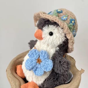 K&ouml;nnte beinhalten: Ein kleiner Pl&uuml;schpinguin, der einen braunen geh&auml;kelten Hut mit blauen Blumen tr&auml;gt. Der Pinguin h&auml;lt eine blaue geh&auml;kelte Blume in seiner rechten Flosse. Der Pinguin sitzt in einem braunen Pappeikarton.