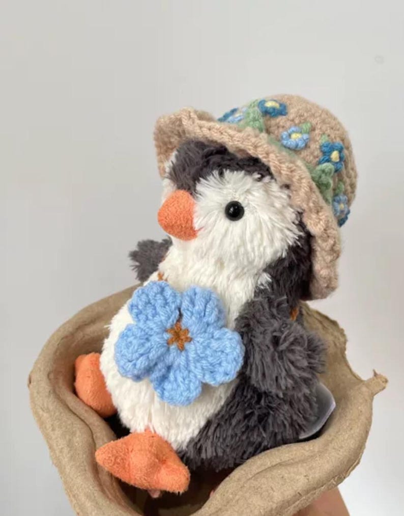 K&ouml;nnte beinhalten: Ein kleiner Pl&uuml;schpinguin, der einen braunen geh&auml;kelten Hut mit blauen Blumen tr&auml;gt. Der Pinguin h&auml;lt eine blaue geh&auml;kelte Blume in seiner rechten Flosse. Der Pinguin sitzt in einem braunen Pappeikarton.