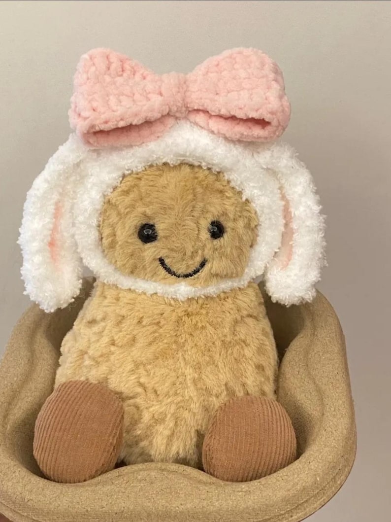 Może przedstawiać: Pluszowa brązowa zabawka z białym puszystym kapeluszem i r&oacute;żową kokardką. Zabawka siedzi w brązowym wiklinowym koszyku.