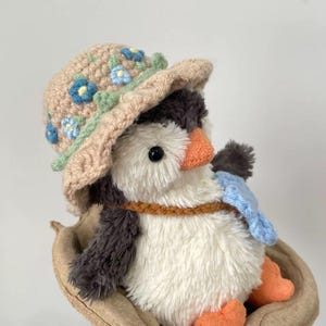 K&ouml;nnte beinhalten: Ein Pl&uuml;schpinguin mit einem geh&auml;kelten Hut mit blauen und gr&uuml;nen Blumen. Der Pinguin sitzt in einem braunen, geh&auml;kelten Nest.