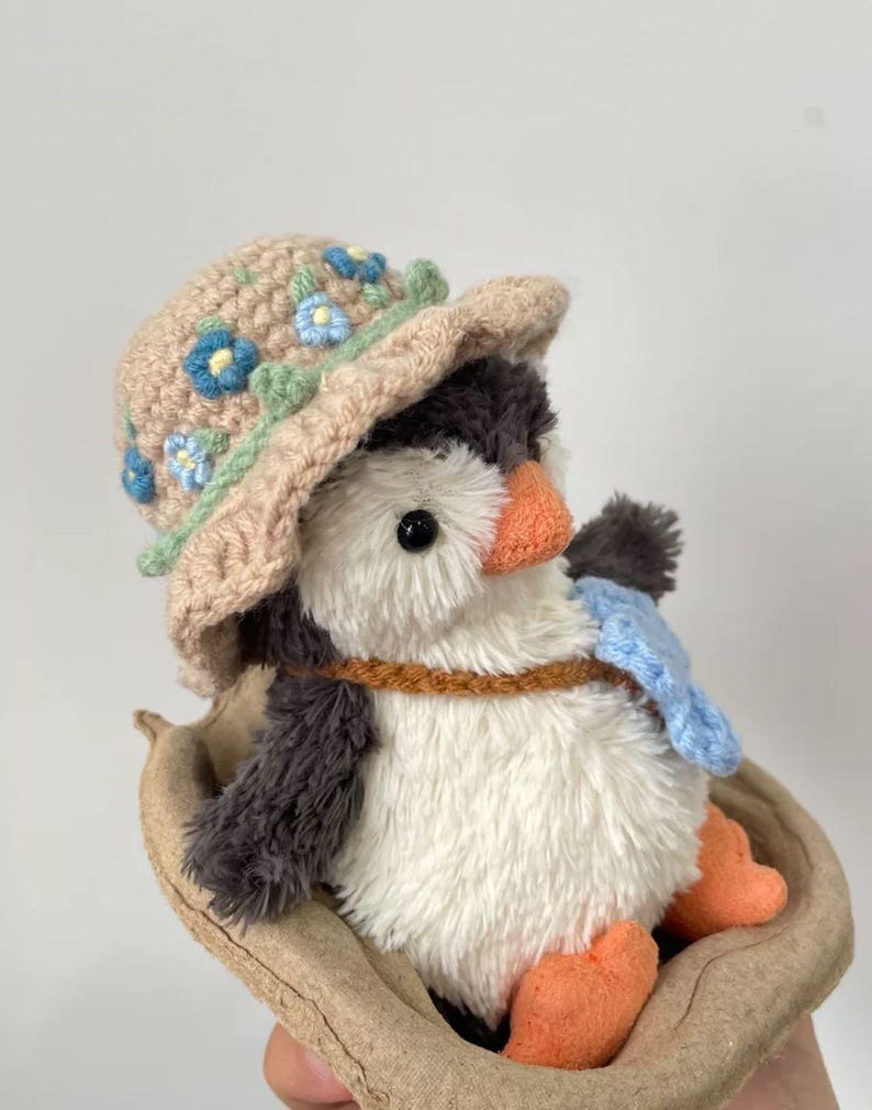 K&ouml;nnte beinhalten: Ein Pl&uuml;schpinguin mit einem geh&auml;kelten Hut mit blauen und gr&uuml;nen Blumen. Der Pinguin sitzt in einem braunen, geh&auml;kelten Nest.